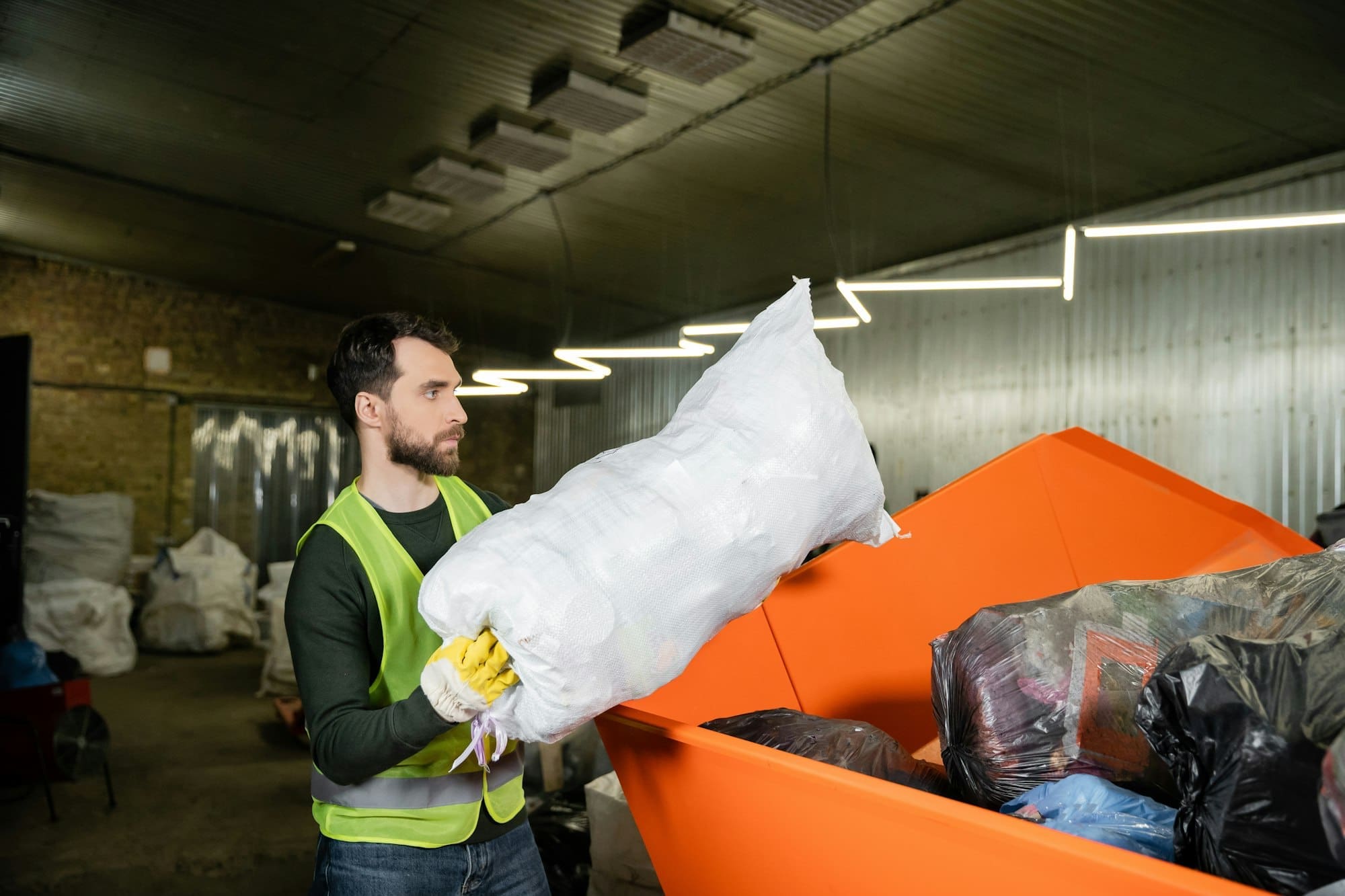 Side view of man in high visibility vest and glove putting sack with trash in container while