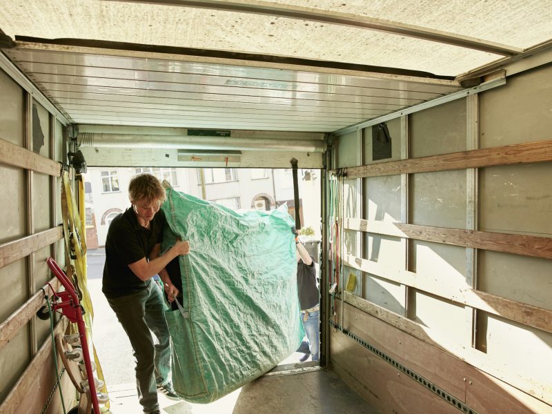 Removals business. A man lifting an item of furniture covered in green plastic into a removals van.